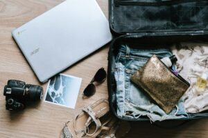 Gut organisiert packen: Ein offener Koffer, daneben Frauensandalen, eine Sonnenbrille, eine Kamera und ein Computer, auf dem sich eine Packliste befindet. Bild: anete-lusina-unsplash