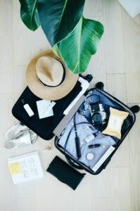 Gut organisiert packen: Ein offener Koffer, daneben unter anderem Frauensandalen, ein Sonnenhut und ein Notizbuch mit einer Packliste. Bild: Pacmarissa-grootes-unsplash