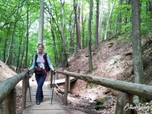 Eine Frau beim Pilgern mit Bauchtasche, Wanderstöcken und Windweste. Bild: Reisen ins Hier